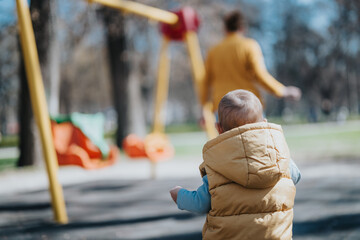A toddler in a yellow jacket explores a park while colorful playground equipment and an adult...
