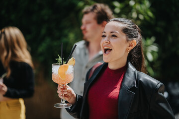 Smiling young woman holding a vibrant cocktail in a cheerful outdoor setting with friends,...