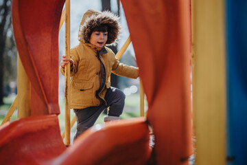 Child dressed warmly with a hooded jacket, playing on a vibrant slide structure in an outdoor park...