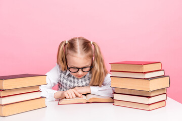 Cute schoolgirl in glasses reading a book surrounded by stacks of book on a pink background,...