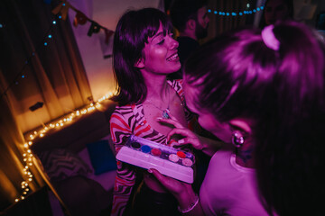 A lively indoor birthday party with friends enjoying face painting under vibrant, colorful...