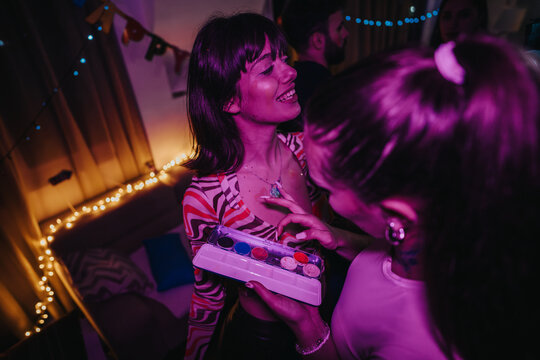 A lively indoor birthday party with friends enjoying face painting under vibrant, colorful lighting. The festive atmosphere is enhanced by decorative lights and cheerful interactions among attendees.