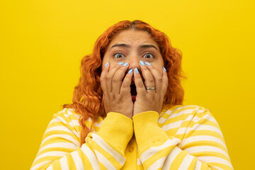 Red haired woman with her hands on her face with a terrified expression on a yellow background with...