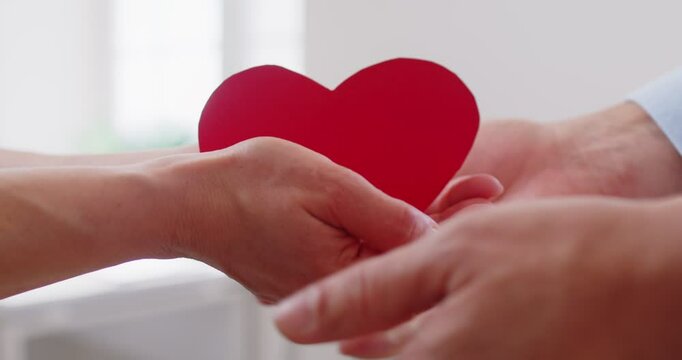 Man tenderly places red paper heart into woman hands, symbolizing their love and bond. Close up of woman and man hands holding red heart together, demonstrating their shared feelings. Love concept.