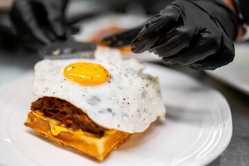 Close-up of a gourmet waffle topped with pulled meat, melted cheese, and a sunny-side-up egg, with...
