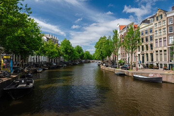 Fototapeta premium Amsterdam, Netherlands – May 21, 2025: Panoramic view of Amsterdam canal, bordered by historical buildings, green trees, and moored boats