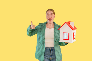 Excited woman holding a miniature house model while giving a thumbs up against a vibrant yellow...