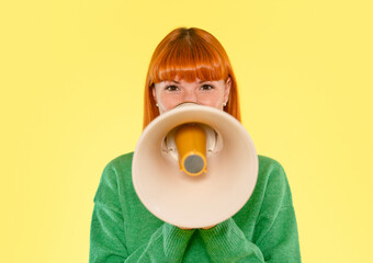 Woman with bright red hair holds a megaphone in front of a yellow background while wearing a green sweater, conveying a message or call to action