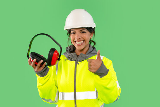 Woman in bright safety gear smiling while holding protective headphones and giving a thumbs up in a vibrant green background - Powered by Adobe