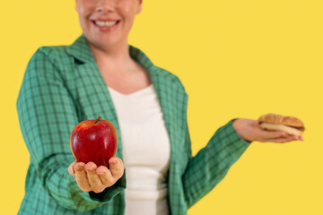 Woman holding an apple in one hand and a sandwich in the other against a bright yellow background,...
