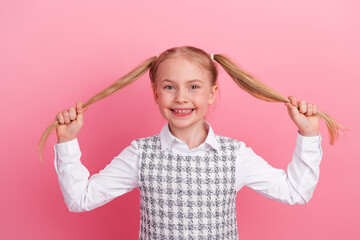 Cheerful schoolgirl holding pigtails against pink background wearing school uniform expressing happiness and playfulness