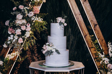A sleek three-tier wedding cake with a smooth gray finish, adorned with pastel pink and white flowers and eucalyptus leaves. Displayed on a geometric stand surrounded by a floral arch and wooden