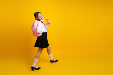 Happy schoolgirl with pink backpack waving on yellow background, showcasing education and...
