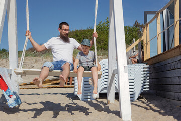 A bearded father with his son swings on a beach swings