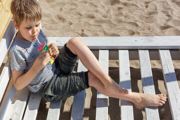 A cute little boy sits on a wooden beach chair and drinks juice from a small packet