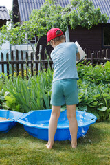 A little boy in a red hat is photographed from behind pouring water into a small pool