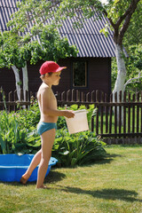 A little boy with a red hat and a white bucket in his hands is getting ready to cool off in the pool in his grandmother's garden on a hot summer day