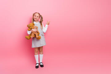 Smiling schoolgirl wearing a stylish uniform holding a teddy bear on a pink background showing a...