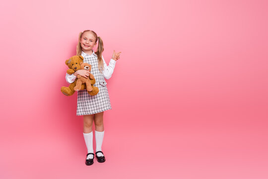 Smiling schoolgirl wearing a stylish uniform holding a teddy bear on a pink background showing a peace sign