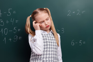 Thoughtful young girl in school uniform solving math equations on green chalkboard, representing education and childhood learning