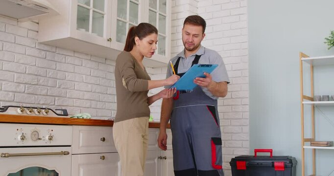 Man plumber talking with client signing contract for plumbing services at home. Male mechanic or repairman consulting woman customer standing in kitchen. Maintenance and repair service. 4k video.
