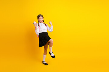 Fototapeta na wymiar Cheerful schoolgirl holding a phone and posing on a yellow background in stylish uniform with a backpack