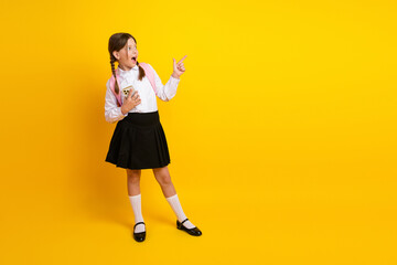 Joyful schoolgirl holding smartphone pointing with finger on a yellow background concept of...