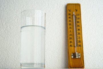 Glass of water in focus with blurred wooden thermometer in background. Concept of hydration, indoor...