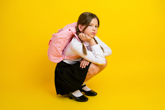 Schoolgirl sitting against bright yellow background wearing a school uniform and pink backpack, looking thoughtful and stylish