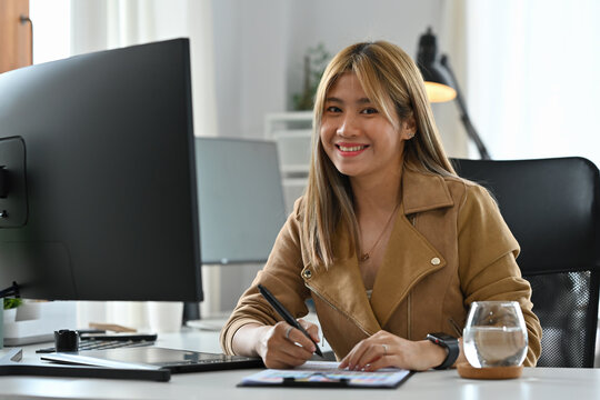 A smiling female professional designer sitting at her desk, surrounded by design tools, looking cheerful and confident
