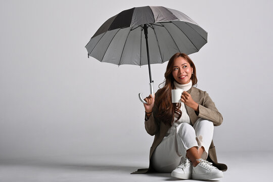Studio shot of a smiling young Asian businesswoman holding an open umbrella and a coffee cup over a clean backdrop, Trust and protection, ideal for insurance or financial service advertisements