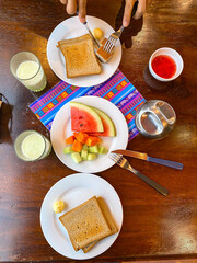 Healthy breakfast in hotel concept. Simple meal of fresh fruits, toasted bread, fruit juice. Person cutting food on plate. Top down angle flat lay of breakfast setting, wooden table. La Paz, Bolivia.