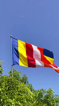 : A vibrant Buddhist flag waving in the wind with the moon and a bird captured in the clear blue daytime sky above.