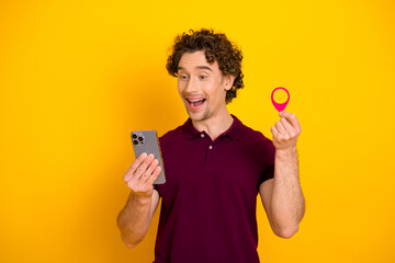 Happy man in a burgundy polo shirt holding a smartphone and a pink locator tag while smiling...