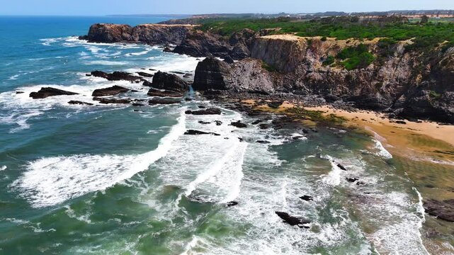 Wide aerial drone shot moving over large rolling waves and a rugged rocky beach with towering cliffs at Praia da Am&aacute;lia, Costa Vicentina, Portugal. Showcasing the powerful ocean meeting dramatic coast