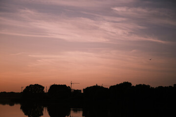 Construction cranes and urban buildings silhouette at sunset, reflected in lake with nature in...
