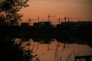 Construction cranes and urban buildings silhouette at sunset, reflected in lake with nature in...