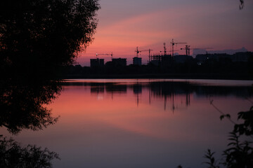 Construction cranes and urban buildings silhouette at sunset, reflected in lake with nature in foreground, evening skyline with industrial development. Contrast between nature and construction