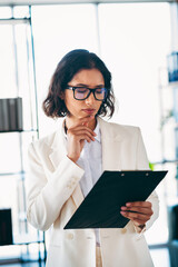 Charming businesswoman analyzing documents in a modern office environment with a thoughtful and...