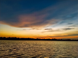 Majestic sunset scene featuring warm golden tones over water and silhouetted trees in the foreground, transitioning to cooler blue hues in the upper sky. Ideal for nature themed background