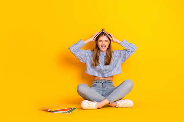 Young student sitting with a book on her head, having fun, laughing against a vibrant yellow...
