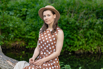 a woman sitting on a fallen tree by the river. She is wearing a brown dress with white polka dots...