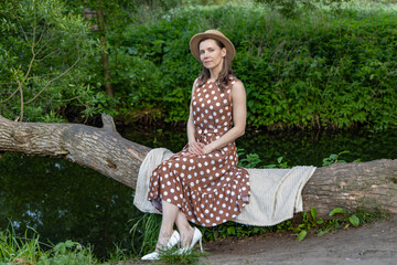 a woman sitting on a fallen tree by the river. She is wearing a brown dress with white polka dots...