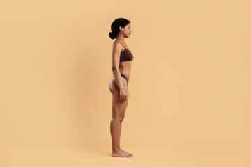 Side profile of young African American woman in underwear standing upright on beige background,...