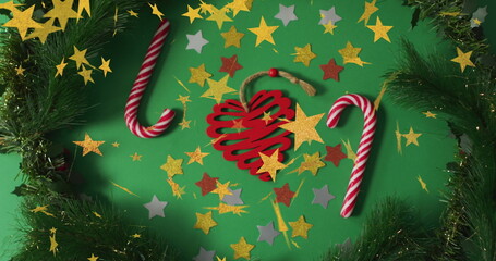 Red wooden heart ornament lying on green table, with candy canes, pine garland, star confetti