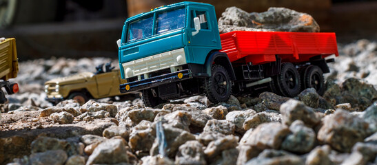 A teal and red toy dump truck navigates a rocky miniature landscape, hinting at imaginative construction play with blurred toy vehicles in the backdrop.