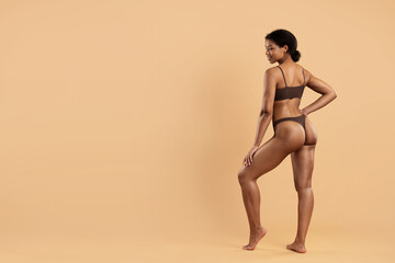 Full body view of fit African American woman in brown underwear posing confidently with smile,...