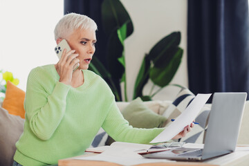 Confident woman with white hair on phone, reviewing documents at home with laptop, expressing focus...