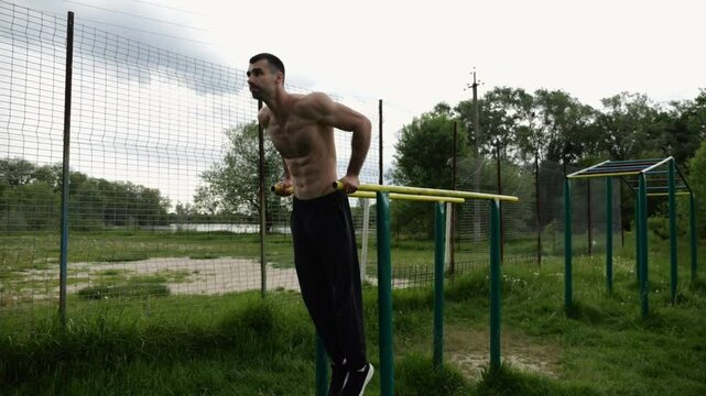 Young sportsman doing push ups on parallel bars at nature. Strong sporty man exercising at sports ground. Male shirtless athlete training hard outdoor. Concept of active sportive lifestyle. Slow mo