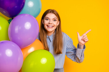 Fototapeta premium Cheerful young woman pointing and posing with colorful balloons against a vibrant yellow background in casual attire